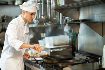 Young man restaurant employee is busy preparing dish in kitchen. Worker roasts food in frying pan, cooks soup and performs auxiliary tasks..
