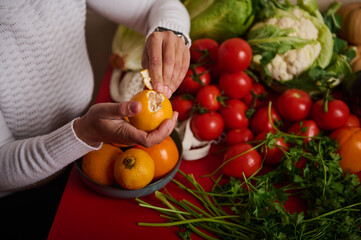 Person Peeling Orange Among Fresh Tomatoes, Herbs, and Vegetables in Bright Kitchen