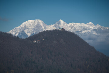 Snow-covered peaks of the Kamnik&ndash;Savinja Alps behind the forested hill of Smarna gora, Slovenia, on a clear winter day with bright blue sky and soft clouds.