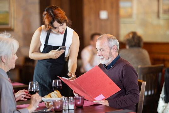 Young woman waitress takes order from couple elderly man and elderly woman in restaurant