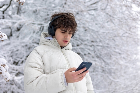 Teenager Listening to Music on Headphones and Using Smartphone Outdoors in Snowy Winter Park.Young Man in Headphones Holding Smartphone in Frosty Forest — Winter Lifestyle and Digital Mood. Copyspace