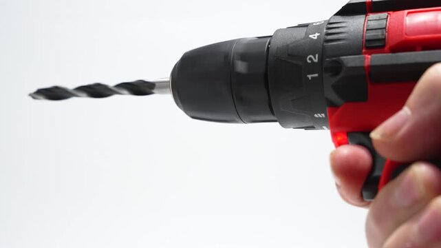 Close-up cordless drill driver with twist drill bit rotates on a white background. A hand grips the trigger, showing the chuck and torque ring in studio light.
