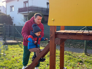 A father assisting his young son on a playground structure, with a yellow panel in the background, in a sunny outdoor setting