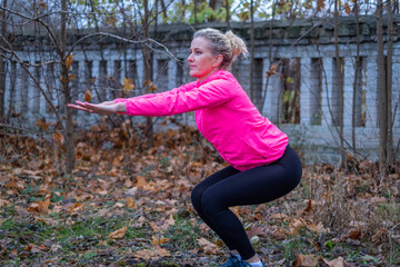 Woman with curly hair in a pink jacket performs squats outdoors among fallen leaves, showcasing a healthy lifestyle and fitness movement in a seasonal setting