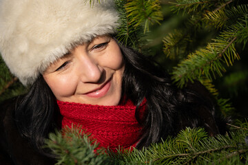 Mid-age woman with a joyful smile, wearing a white hat and red scarf, surrounded by evergreen branches, capturing the essence of beauty and happiness in nature