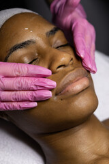 Naklejka premium African woman in beauty clinic doing a facial evaluation and a cosmetology treatment. Woman on an esthetician appointment of facial. use mask do skincare routine