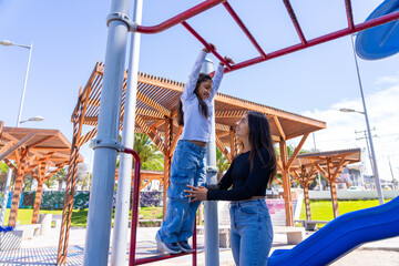 Mother supporting her daughter swinging on playground monkey bars, having fun outdoors