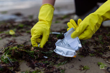 Hands in yellow gloves picking up crushed packaging and litter during beach cleanup, showing pollution, environmental damage and the importance of eco awareness and coastal protection.