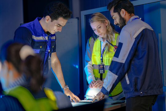 A diverse team of engineers, a Caucasian woman in a safety vest with Indian and Caucasian male colleagues, collaborates late at night. They are focused, reviewing plans in a factory control room. - Powered by Adobe