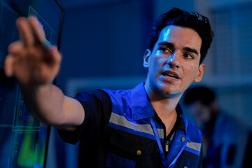 A young, happy Caucasian male engineer in a uniform smiles while using a stylus on a screen. He is working in a high tech control room or factory at night with his team.