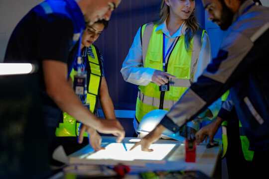n Indian engineer uses a radio for incident response in a smart factory. His female colleague in a safety vest checks AI system data on a glowing panel in the command center.