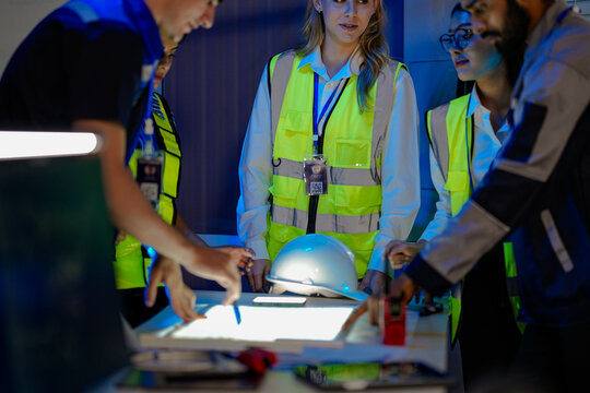 n Indian engineer uses a radio for incident response in a smart factory. His female colleague in a safety vest checks AI system data on a glowing panel in the command center.