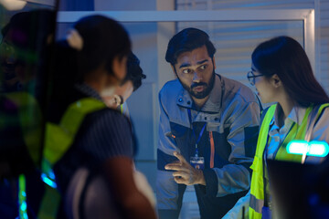 An Indian male engineer in a uniform speaks to his female colleagues. The diverse team, including an Asian woman in a safety vest, collaborates in a control room at night.