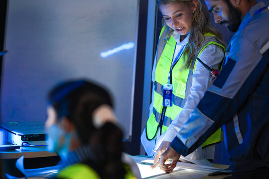 A logistics management team reviews supply chain blueprints on a light table. The diverse group, led by a woman in a safety vest, plans an AI strategy for optimization.
