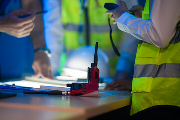 A close up of an engineering team collaborating at night. Hands point at a glowing blueprint on a light table, with a hardhat and walkie talkie on the desk.