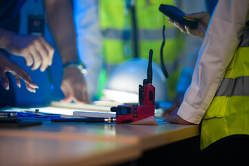 A close up of an engineering team collaborating at night. Hands point at a glowing blueprint on a light table, with a hardhat and walkie talkie on the desk.
