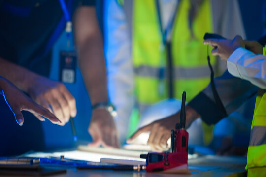 A close up of an engineering team collaborating at night. Hands point at a glowing blueprint on a light table, with a hardhat and walkie talkie on the desk.