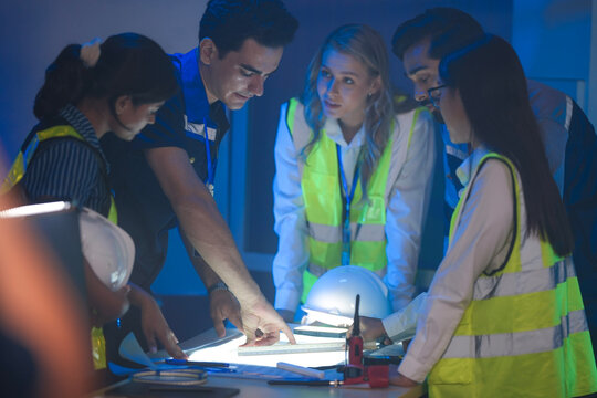 A large, diverse team of engineers in safety vests collaborates at night. An Indian woman holds a hardhat as the team reviews plans in a factory control room.