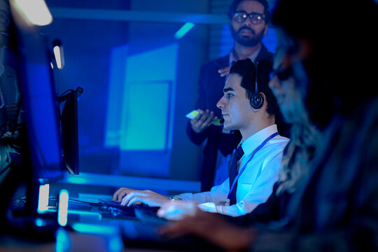 IT support team lead oversees a junior agent handling a complex technical issue. He offers real time advice while reviewing progress on screen in a busy operations center.