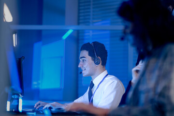 Dynamic night shift collaboration. Male manager provides guidance to a focused male colleague wearing a headset at his computer in a modern tech office during an intense work session.