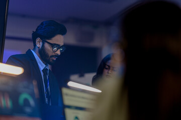 Focused Indian businessman in glasses analyzing financial data charts on night shift in blue-lit modern office, working on urgent corporate project with team.