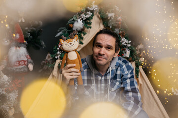 A smiling, dark-haired man with wrinkles on his face, wearing jeans and a shirt, sits near a...