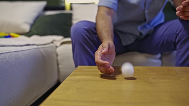 A person playfully bounces a white ping pong ball on a wooden coffee table in a home setting. The engaging sound adds to the atmosphere of simple indoor fun and relaxation.