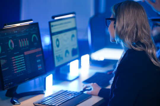 Focused Indian male operator in glasses and headset discussing data with a colleague in a blue-lit control room, managing urgent network operations or customer support.