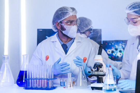 Diverse team of scientists in protective gear collaborating in modern blue lab, analyzing DNA helix structure on monitor and testing chemical samples for medical research.