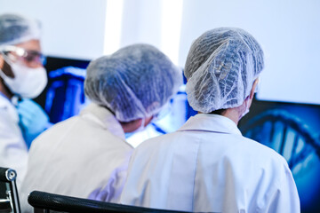 Fototapeta premium Diverse team of scientists in protective gear collaborating in modern blue lab, analyzing DNA helix structure on monitor and testing chemical samples for medical research.