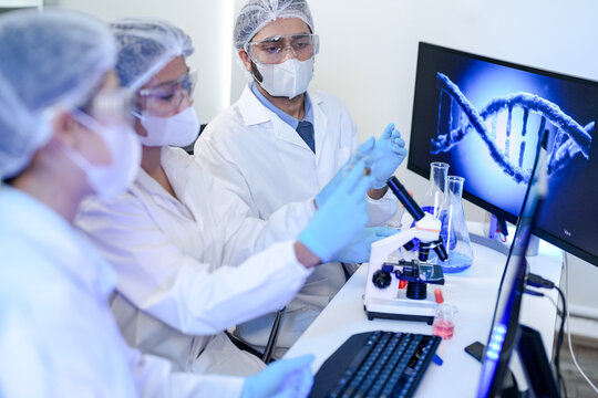 Diverse team of scientists in protective gear collaborating in modern blue lab, analyzing DNA helix structure on monitor and testing chemical samples for medical research.