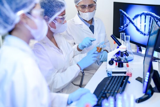 Diverse team of scientists in protective gear collaborating in modern blue lab, analyzing DNA helix structure on monitor and testing chemical samples for medical research.