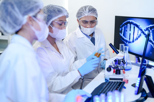 Diverse team of scientists in protective gear collaborating in modern blue lab, analyzing DNA helix structure on monitor and testing chemical samples for medical research.