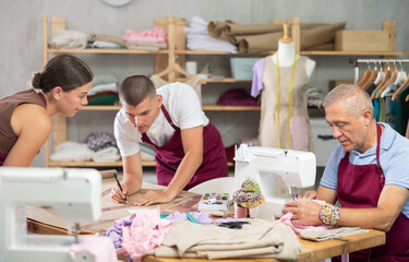 Working day in sewing workshop, textile industry. Senior man master sew clothes in sewing machine, guy and girl apprentice helper draw perform paper prototype of product parts