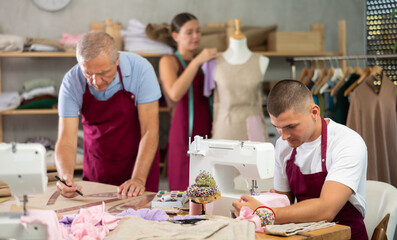 Young tailor sews clothes on a machine next to a mature worker who draws pattern. Tailors sew...