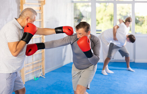 Man with old man at group boxing lesson are learning new technique of body blowing, mam is sparring with guy in background