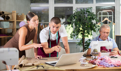 Female fashion designer with a team of tailors designs and sews clothes. Elderly tailor sews next to the employees of the atelier. Dressmaker shows a sketch of clothes on a laptop
