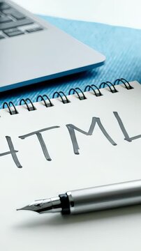 Close up photograph featuring a notepad with the handwritten acronym HTML on its pages. A sleek silver fountain pen is placed on top of the notepad, with a laptop visible in the background