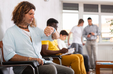 Man sitting in lobby of medical facility and looking impatiently at watch while waiting for doctor...