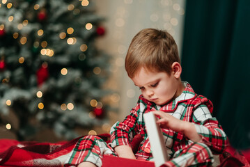 child in festive plaid pajamas opening a present while sitting near a beautifully lit Christmas tree. Warm holiday atmosphere with soft bokeh lights and cozy seasonal colors.