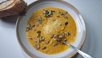 Creamy pumpkin soup topped with seeds and grated cheese is served with a slice of crusty bread. This image conveys the concept of warm homemade comfort food and cozy autumn dining