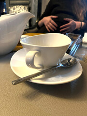 A delicate white tea cup sits on a saucer with a spoon beside it, while a person relaxes in a cozy corner of the cafe, enjoying the atmosphere and warmth