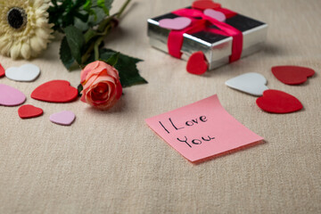 Valentine’s Day composition featuring a handwritten ‘I love you’ note on a soft fabric surface, decorated with heart-shaped cutouts, a rose, flowers, and a wrapped gift. Ideal for romantic themes, cel