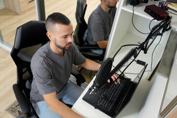 In a home studio, a man focuses on recording a song using headphones and a microphone connected to his laptop while surrounded by modern equipment in a bright room