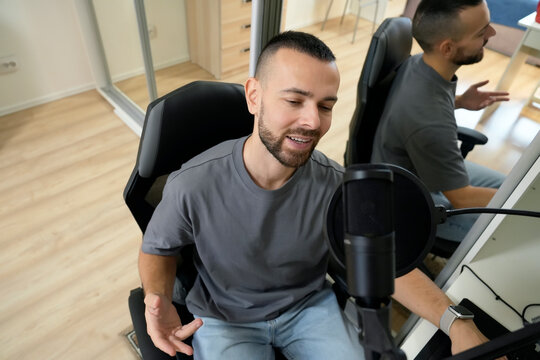 A man sits in a chair, recording a song with a microphone in front of him. He wears headphones and uses a laptop for streaming. This intimate home setup showcases creativity and communication