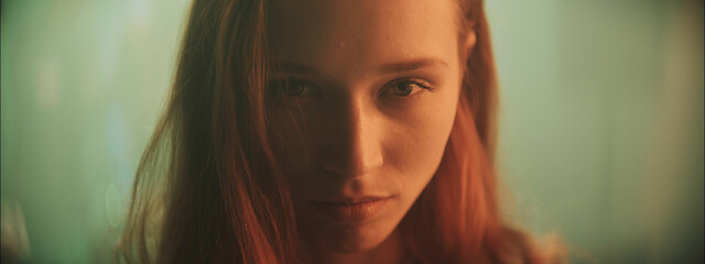Young woman with red hair gazes intensely at the camera in a softly lit setting at dusk