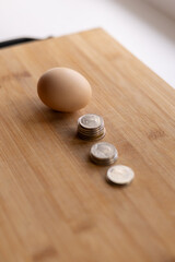 A brown egg sits on a wooden surface beside three small stacks of coins, illustrating a simple concept of cost, value, or price comparison in soft natural light.