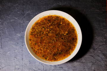 A close-up of a bowl of hot golden broth with visible oil droplets, herbs, and spices floating on the surface. Warm, homemade soup with rich color and appetizing texture.