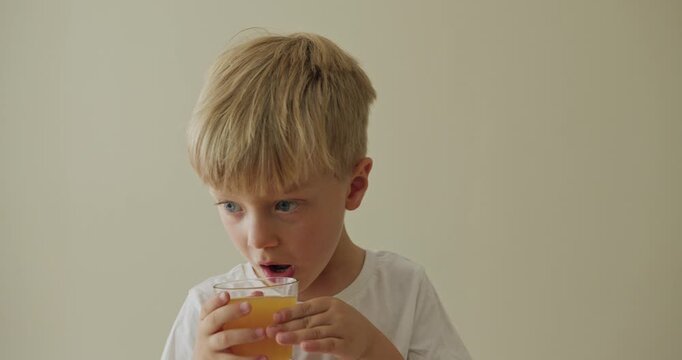 oy with blond hair in a white T-shirt thoughtfully drinks orange juice and looks out the window.