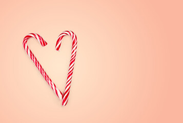 Two red and white striped candy canes are positioned to form a heart shape against a soft pink background. This festive decoration symbolizes the holiday spirit.